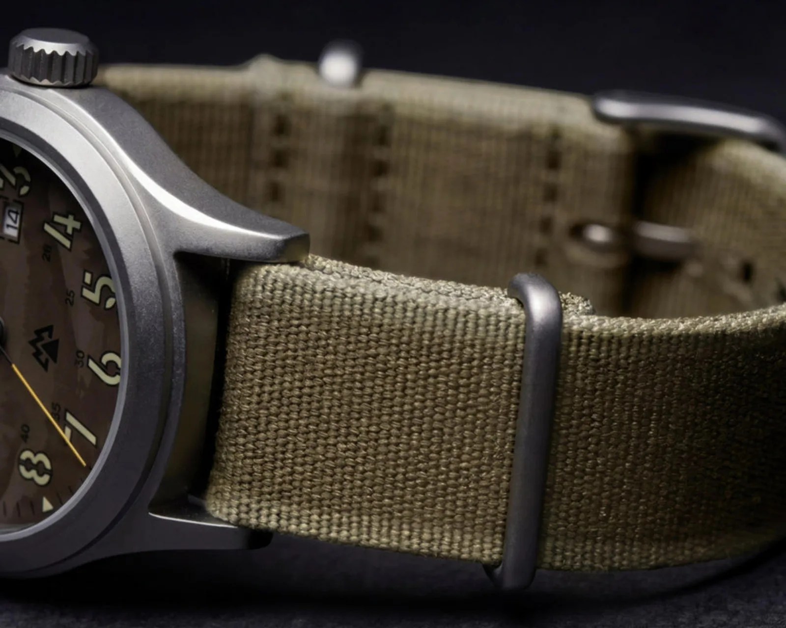 Close-up of a wristwatch with a matte metal case and olive green woven fabric strap with metal loops.