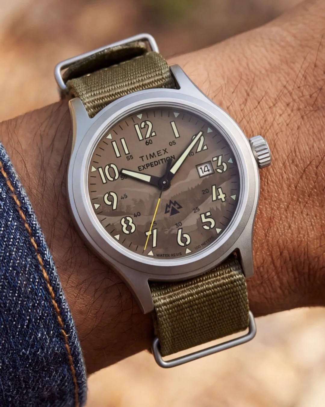 Timex Expedition wristwatch with a beige dial, luminous numerals, date window, and olive green fabric strap worn on a wrist.