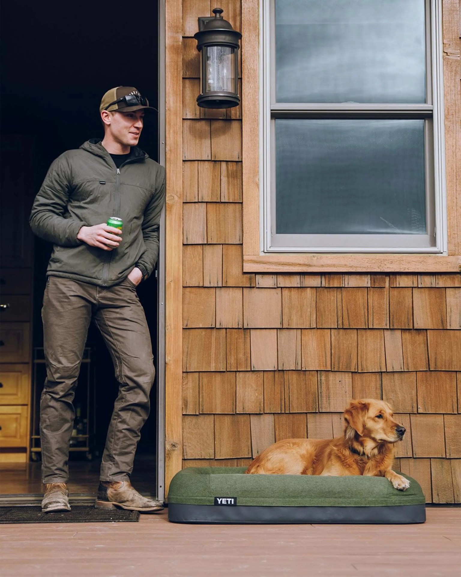 Golden retriever lying on a green YETI dog bed on a wooden porch next to a man in outdoor clothing.