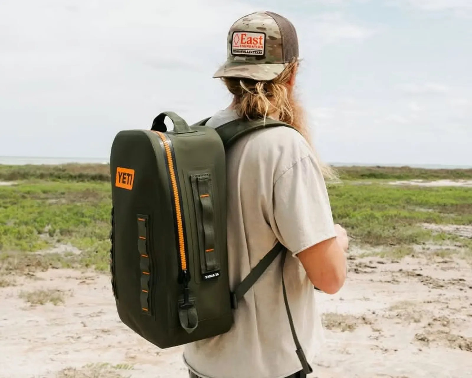 Olive green YETI backpack with orange zipper worn by a person in a beige shirt and camo hat outdoors.