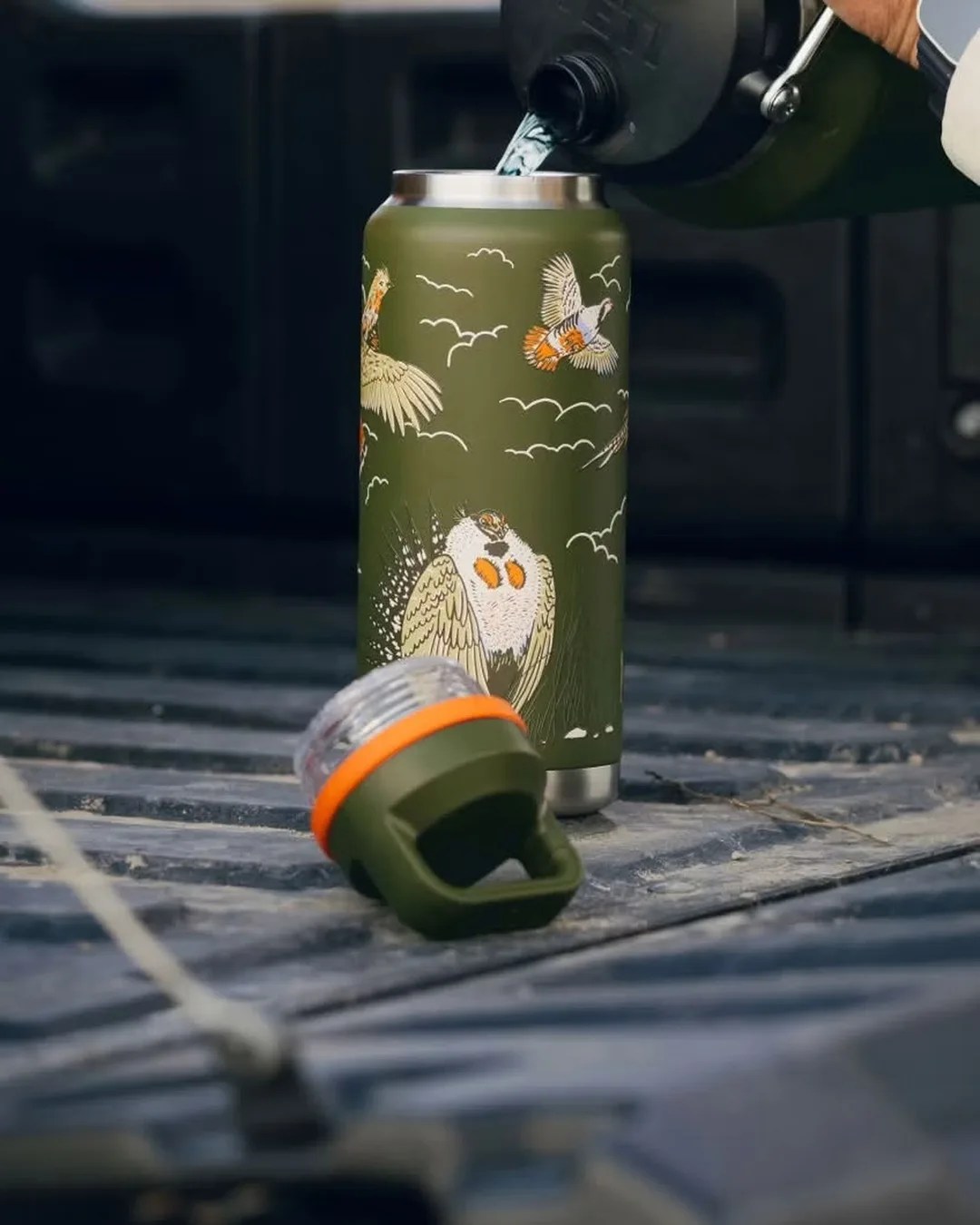 Green insulated bottle with bird illustrations and a detachable lid with an orange ring on a truck bed.